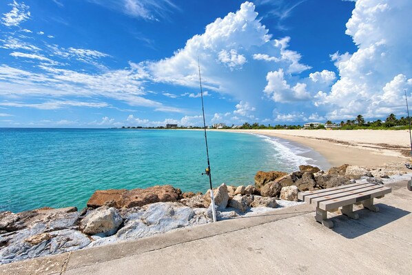 A beach with clear turquoise water, a rocky shoreline, and a concrete walkway with a bench.