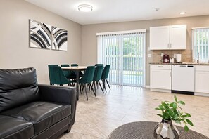 Un salon moderne avec un canapé en cuir noir, un coin repas avec des chaises vertes et une cuisine avec des meubles blancs.