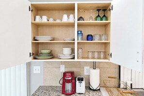A kitchen with open cabinets displaying dishes, glasses, and jars. Below, there is a coffee maker, toaster, and a water dispenser.