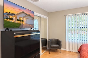 A modern bedroom with a TV on a stand, a bed, a chair, and a window with blinds.