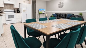 A modern kitchen with white appliances, a dining area with teal chairs and a wooden table, and a living area with a black leather sofa. - Centrally located 4/2 Home with a private backyard available for Longer Stays! (Port St. Lucie)