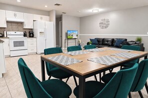 A modern kitchen with white appliances, a dining area with teal chairs and a wooden table, and a living area with a black leather sofa. - Centrally located 4/2 Home with a private backyard available for Longer Stays! (Port St. Lucie)