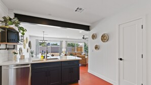 A modern kitchen with dark cabinets, a stainless steel dishwasher, and a white countertop. There is a large window with curtains, a ceiling fan, and a door leading to a living area with a beige sofa and a plant. - Newer, Modern, Work Friendly 2 Bedroom Condo near Downtown PHX (Phoenix)