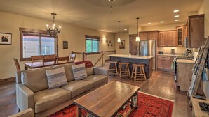 A spacious living room with a sofa, coffee table, and a red rug. There is a kitchen area with wooden cabinets and a bar counter. - Walkable Bigfork Townhome: Fish, Hike & Ski! (Bigfork)