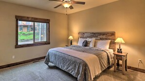 A bedroom with a bed, two bedside lamps, a ceiling fan, and a window with a view of a house and greenery. - Walkable Bigfork Townhome: Fish, Hike & Ski! (Bigfork)