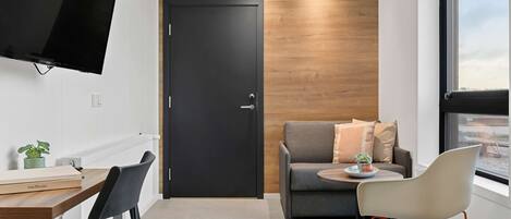 A modern room with a wooden accent wall, a black door, a grey sofa with cushions, a round table with a plant, a TV mounted on the wall, and a desk with chairs.