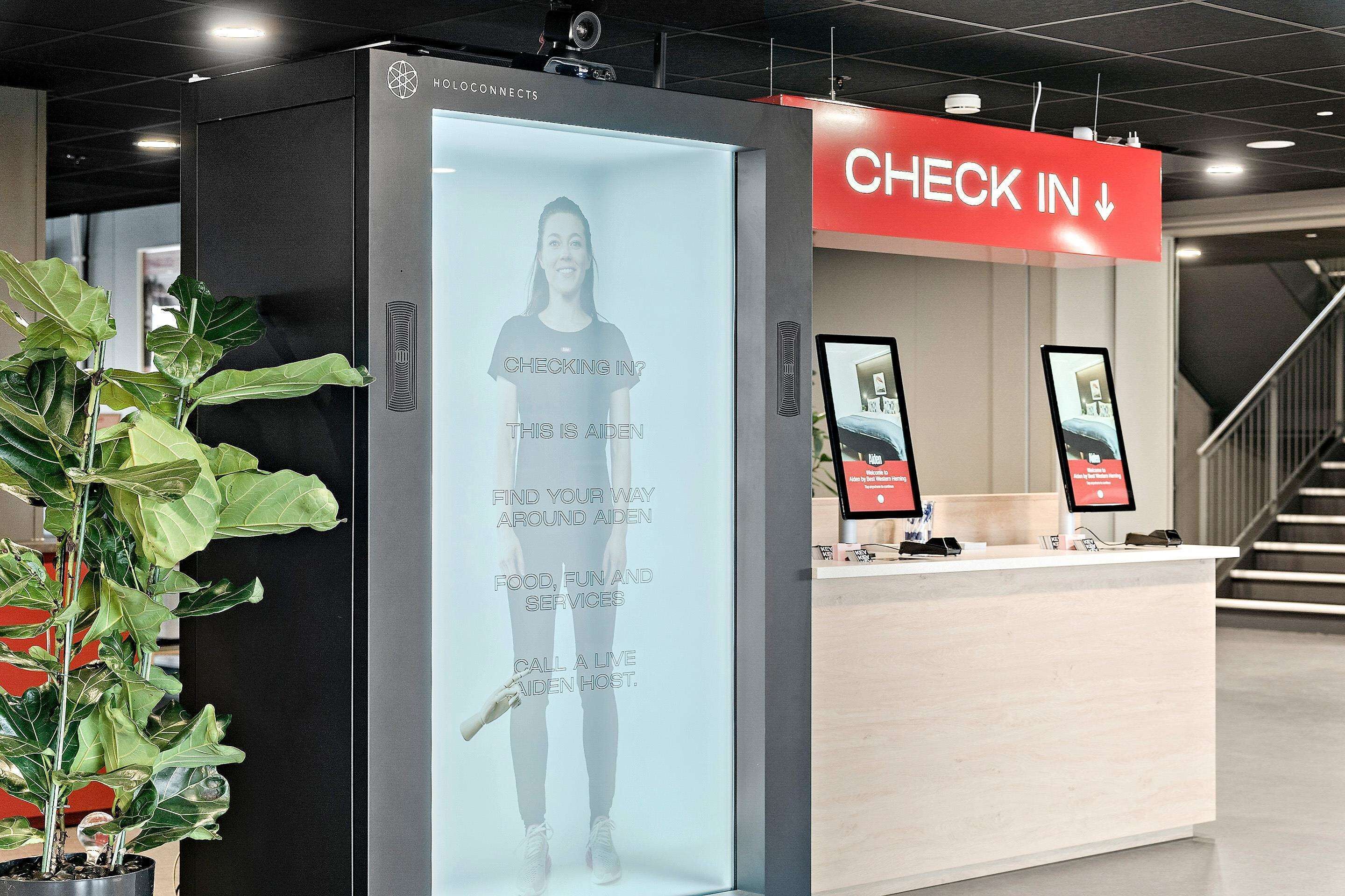 A hotel reception area with a digital screen displaying a welcome message, a 'Check In' sign, and a reception desk with a computer monitor and a tablet.