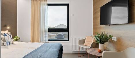 A modern hotel room with a bed, a desk, a chair, a potted plant, and a TV mounted on the wall.
