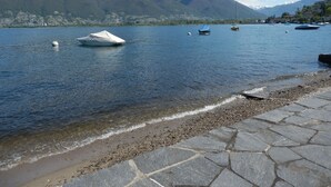 A lake with boats, a pebble beach, and mountains in the background. - Schöne Ferienwohnung App.36 mit Privatstrand (Gambarogno)