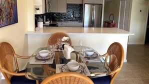A modern kitchen with stainless steel appliances, a dining area with glass tables and wicker chairs, and a large painting on the wall.