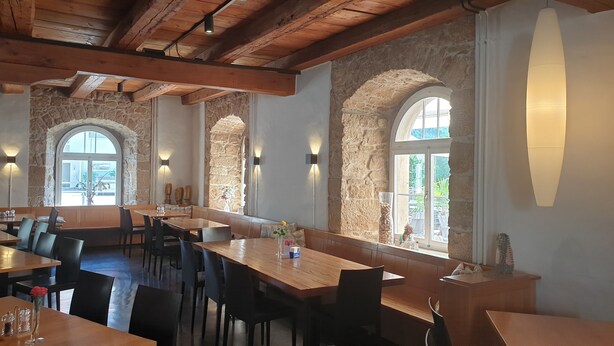 Un restaurante con paredes de piedra, vigas de madera y ventanas de arco. Mesas y sillas dispuestas para comer.