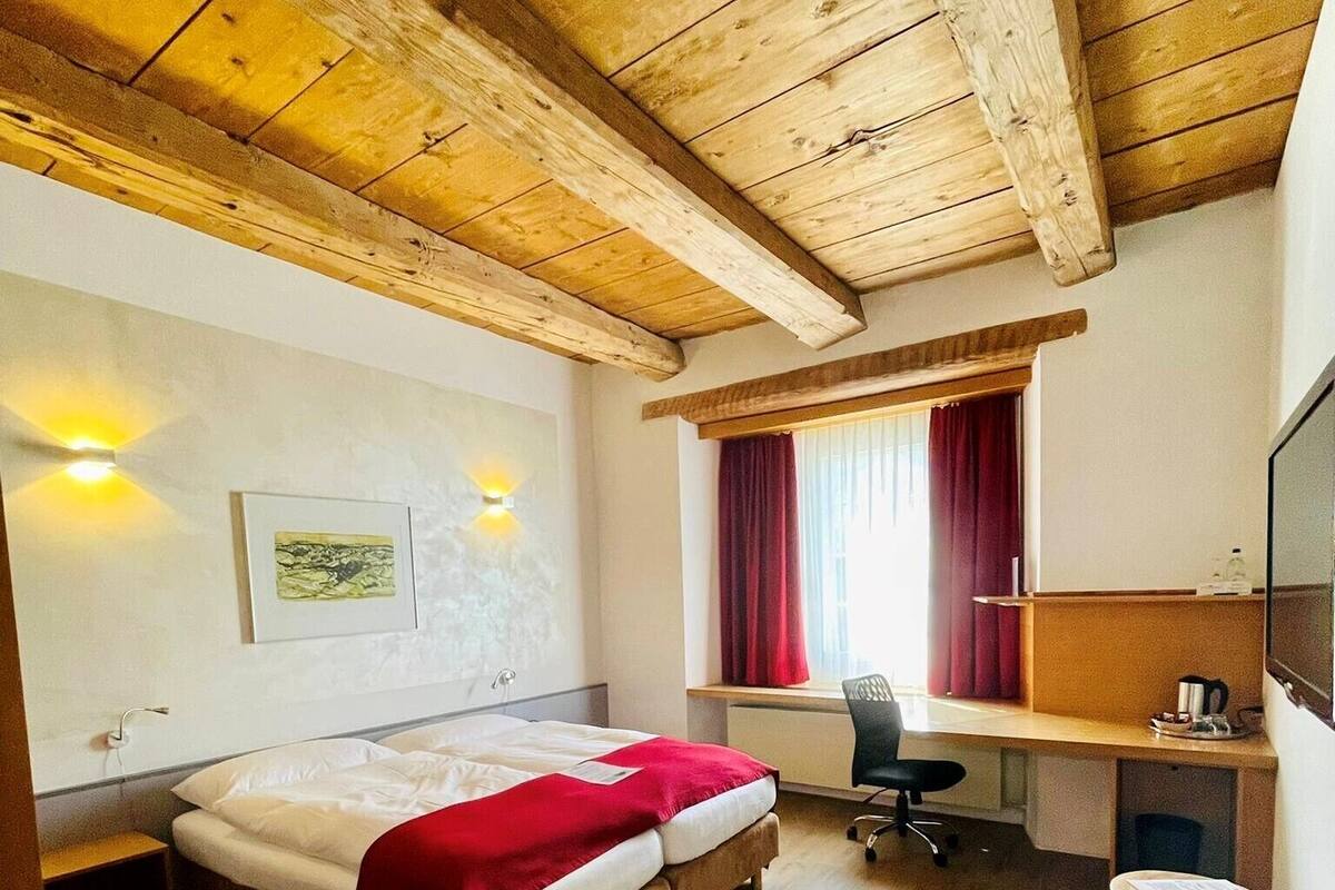 A hotel room with a wooden ceiling, a bed with a red blanket, a desk with a chair, and a window with red curtains.