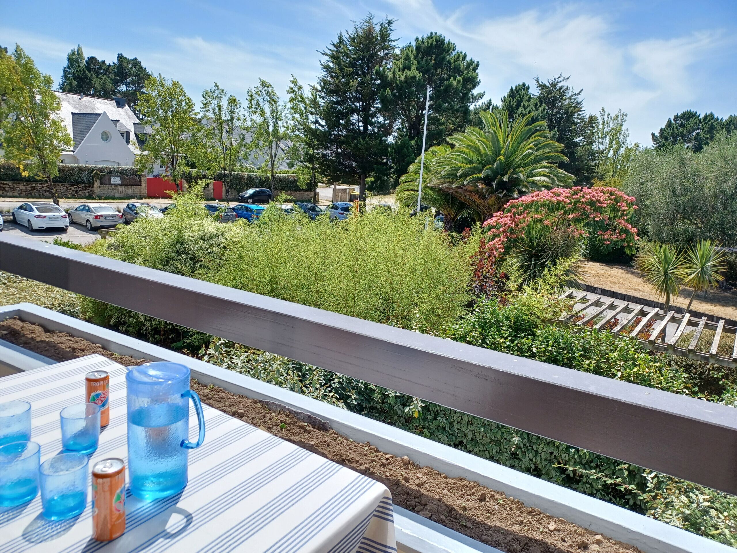 Un balcon avec une table, des boissons et une vue sur un parking et un jardin.