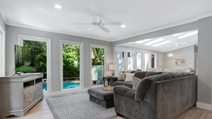 A spacious living room with a grey couch, a flat-screen TV, and a large window overlooking a swimming pool. - 9 Planters Wood Court at The Sea Pines Resort (Hilton Head Island)