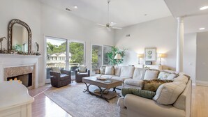 A spacious living room with a fireplace, a ceiling fan, and a large window offering a view of the outdoors. - 13 Spartina Court at The Sea Pines Resort (Hilton Head Island)