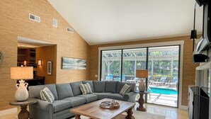Ein Wohnzimmer mit einem grauen Ecksofa, einem Holztisch und einem großen Fenster mit Blick auf einen Pool.