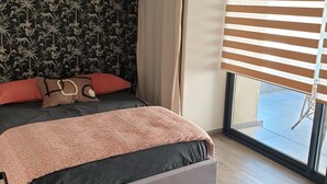 A bedroom with a bed, a window with blinds, a hanging light fixture, and a wall with a tropical pattern. - Mini Villa (Furiani)