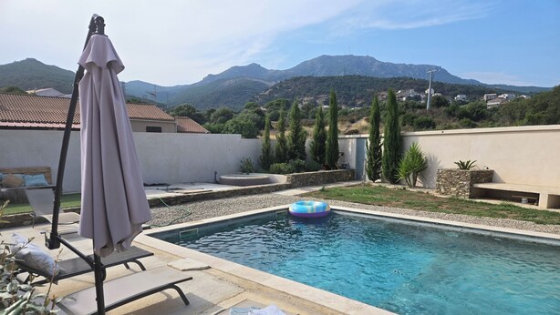 A swimming pool with lounge chairs and an umbrella, surrounded by greenery and mountains in the background. - Mini Villa (Furiani)
