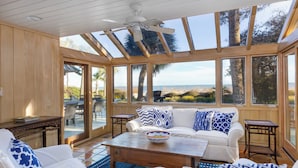 Una veranda soleggiata con un'ampia porta vetrata che si apre su una vista mare, un divano bianco con cuscini a motivi blu e un tavolino da caffè in legno.