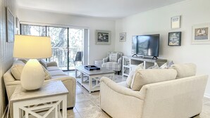 Una sala de estar moderna con un sofá, una mesa de centro y un televisor. Hay un balcón con vistas a árboles.