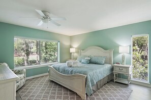 Ein Schlafzimmer mit einem Bett, zwei Nachttischen, einem kleinen Tisch, einem Deckenventilator und großen Fenstern mit Jalousien.