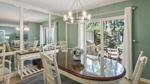 Zona pranzo con tavolo in vetro circondato da sedie bianche, un lampadario e vista sull'esterno.
