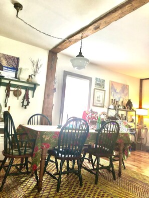 A dining area with a table set for four, surrounded by chairs. A chandelier hangs from the ceiling, and a wooden beam supports the structure.