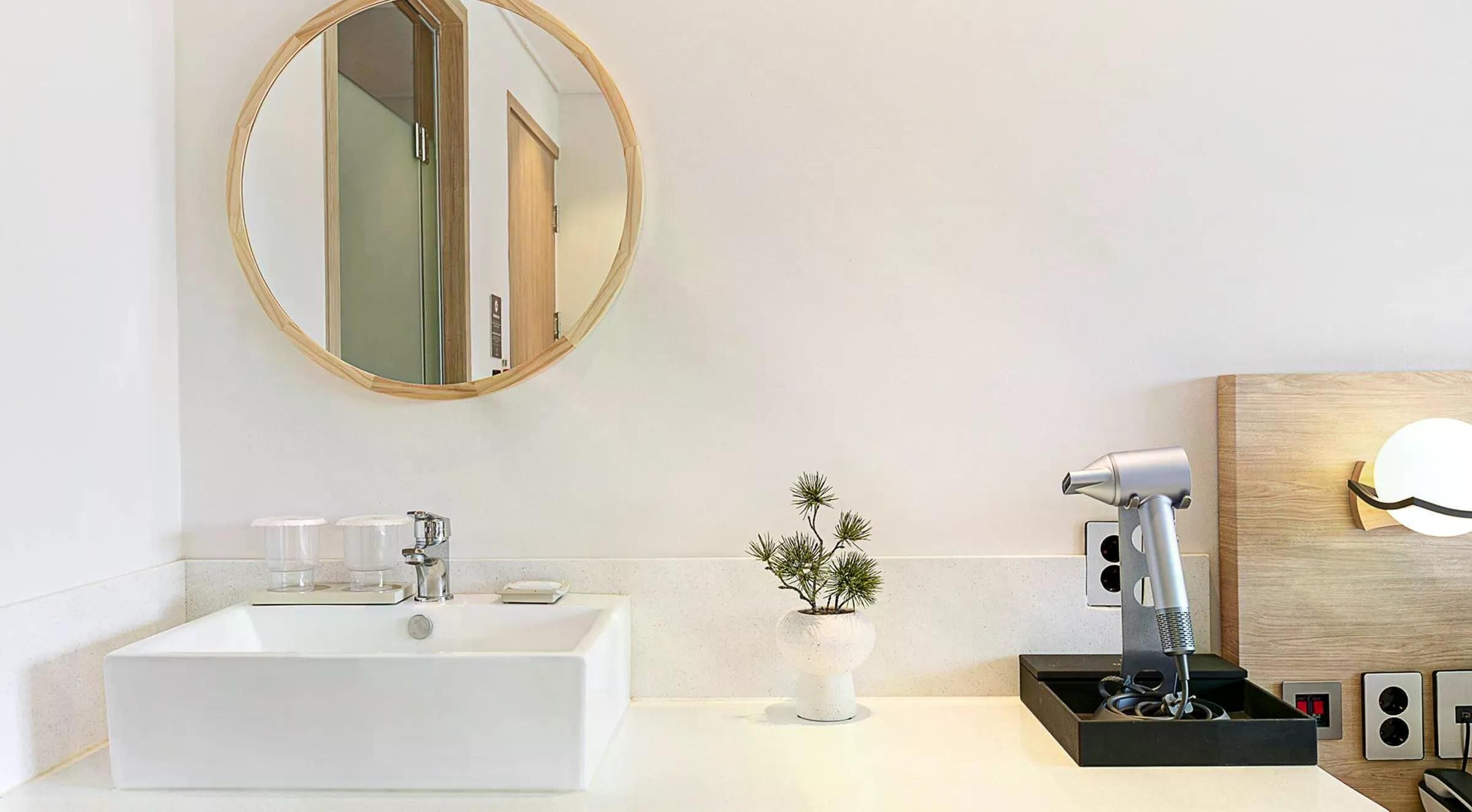 A modern bathroom with a rectangular sink, a round mirror, a small potted plant, and a hairdryer on a wooden panel.