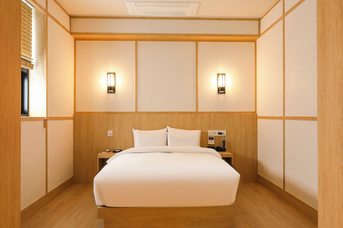 Habitació d'hotel amb un llit gran, dues taules de nit, un llum penjant a la paret i una finestra amb persianes.