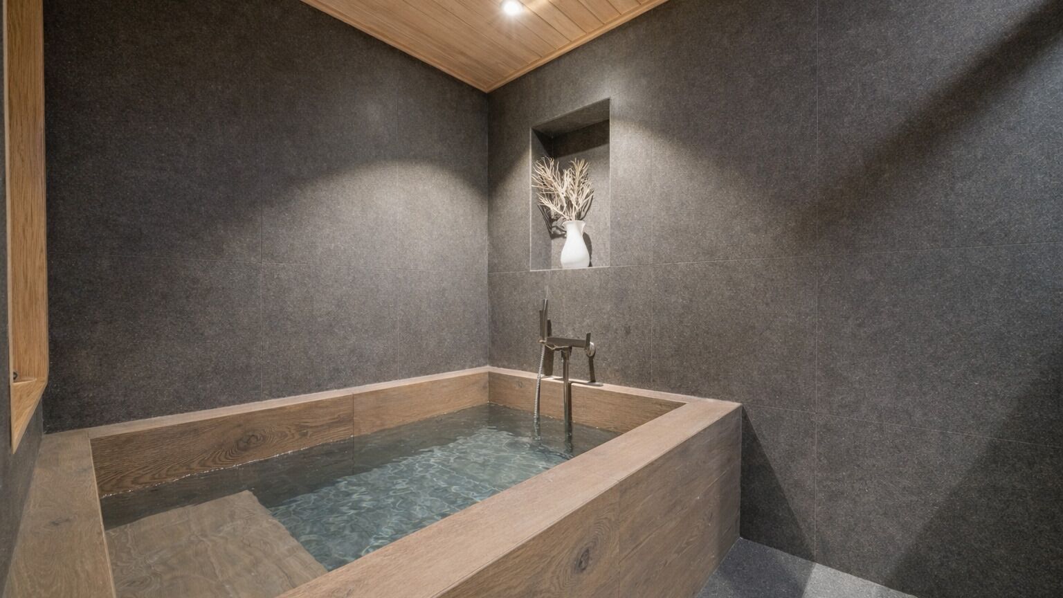 A modern bathroom with a wooden bathtub, dark stone walls, and a small mirror with a vase.