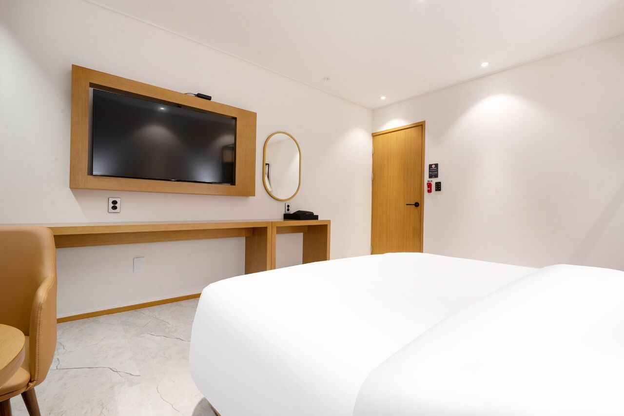 Habitación de hotel con cama, escritorio con computadora, televisor, espejo y puerta con señal de salida de emergencia.