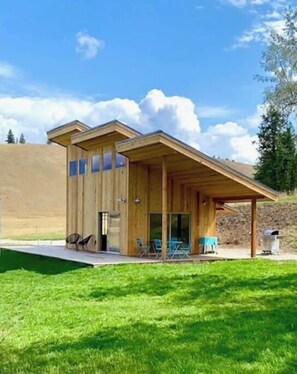 Ein modernes Holzgebäude mit überdachter Terrasse, einer kleinen Sitzgelegenheit im Freien und einem Rasen.