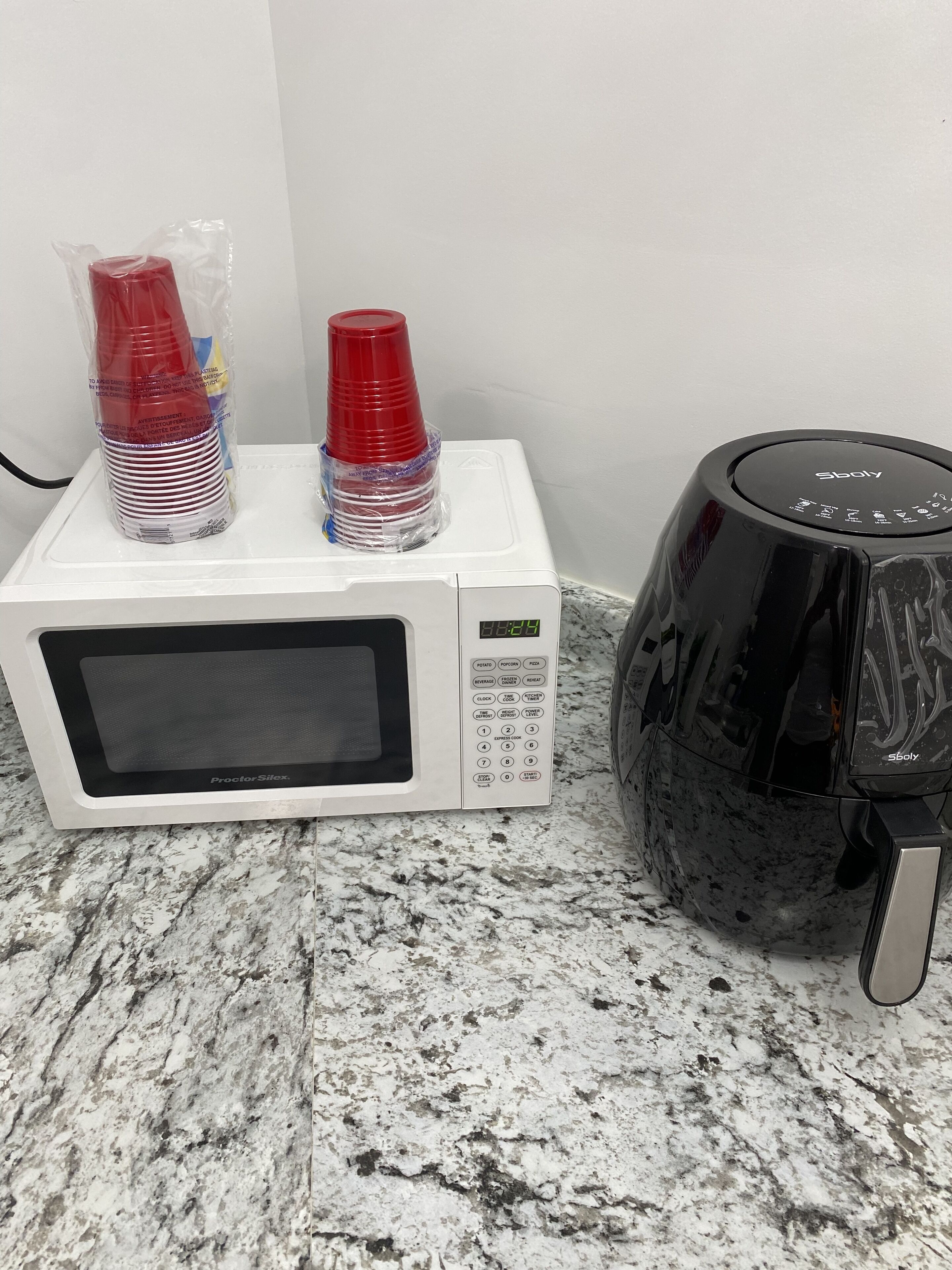 A white microwave oven with a digital display and control panel, a black coffee maker, and stacks of red and clear plastic cups on top.