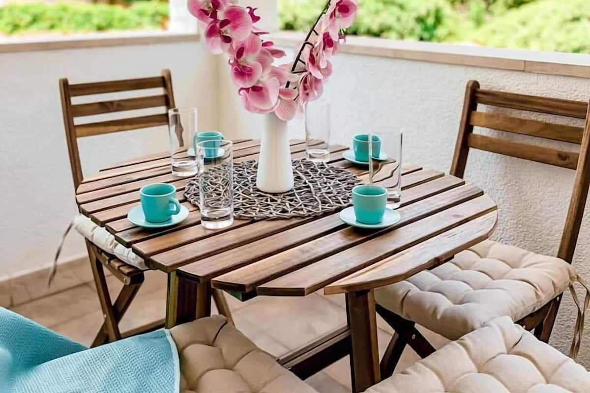 A round wooden table with a white vase of pink flowers, surrounded by four chairs with beige cushions.