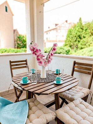 A wooden table with a vase of pink flowers, two glasses, and two cups on a balcony with cushioned chairs.