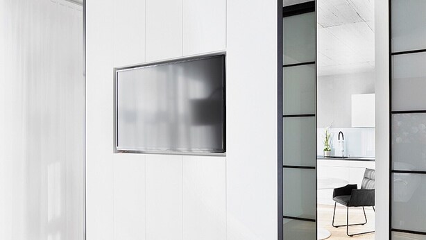 Een modern interieur met een grote flatscreen-tv aan een witte muur, een keukenhoek met een spoelbak en een stoel, en een houten vloer.