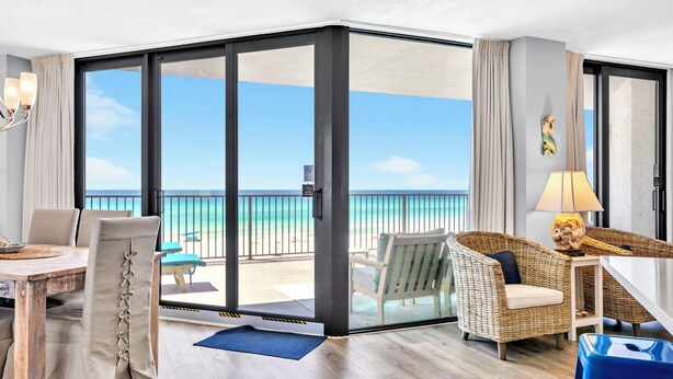 Une chambre d’hôtel dotée d’une porte-fenêtre coulissante donnant sur un balcon avec vue sur l’océan. On y trouve une table à manger avec des chaises, un fauteuil en osier et une petite table avec une lampe.