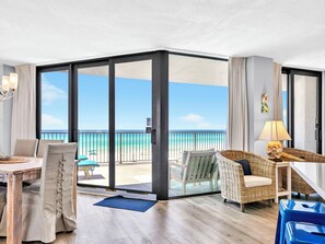 Habitación de hotel con puerta corrediza de vidrio que da a un balcón con vista al océano. Cuenta con una mesa de comedor con sillas, una silla de mimbre y una pequeña mesa con lámpara.