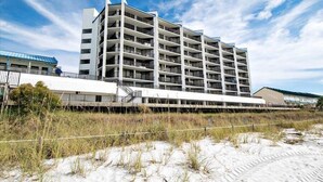 Un hôtel à plusieurs étages, doté d’un toit plat et de balcons, situé près d’une plage de sable où la végétation est clairsemée.