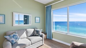 Un salon avec vue sur l’océan, un canapé agrémenté de coussins à motifs et une grande fenêtre avec des rideaux.