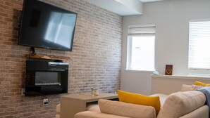 Un salon avec un mur en briques, un téléviseur à écran plat fixé au-dessus d’une cheminée, un canapé beige agrémenté de coussins jaunes et une table basse en bois.