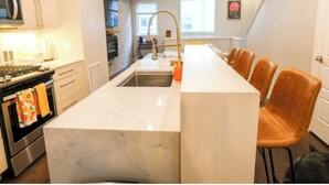 Une cuisine moderne avec un plan de travail en marbre, un four encastré et un évier. Des chaises en bois sont disposées autour d’un îlot central.