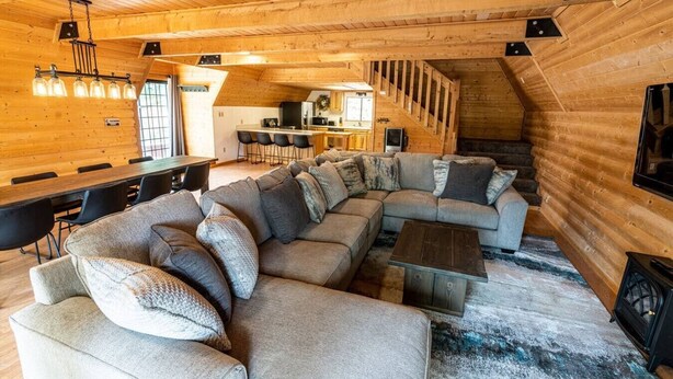 Interior de uma cabana de troncos com um amplo sofá seccional em formato de L, uma área de jantar com uma mesa comprida e uma cozinha ao fundo.
