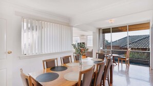 A dining area with a wooden table and chairs, a vase with plants, and a view of a residential area through large windows.