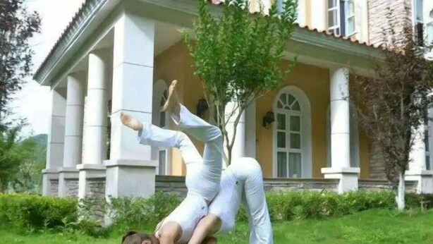 Una persona realizando una postura de yoga sobre césped frente a un edificio con columnas y una ventana.