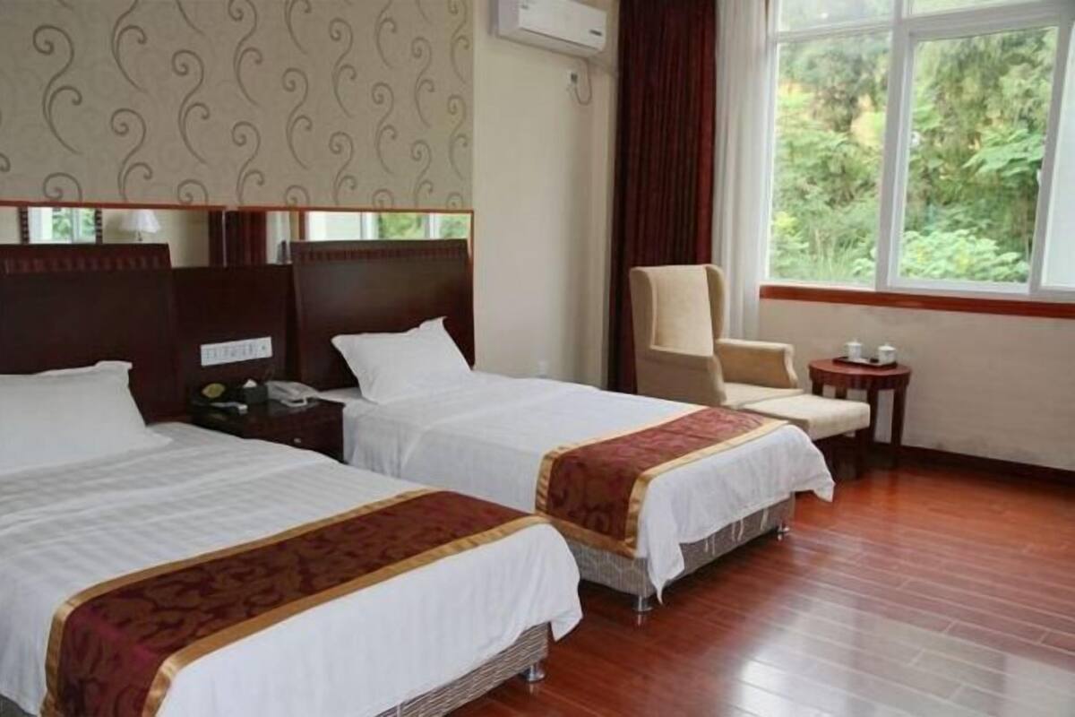 A hotel room with two beds, a chair, a small table, and a window with a view of greenery.