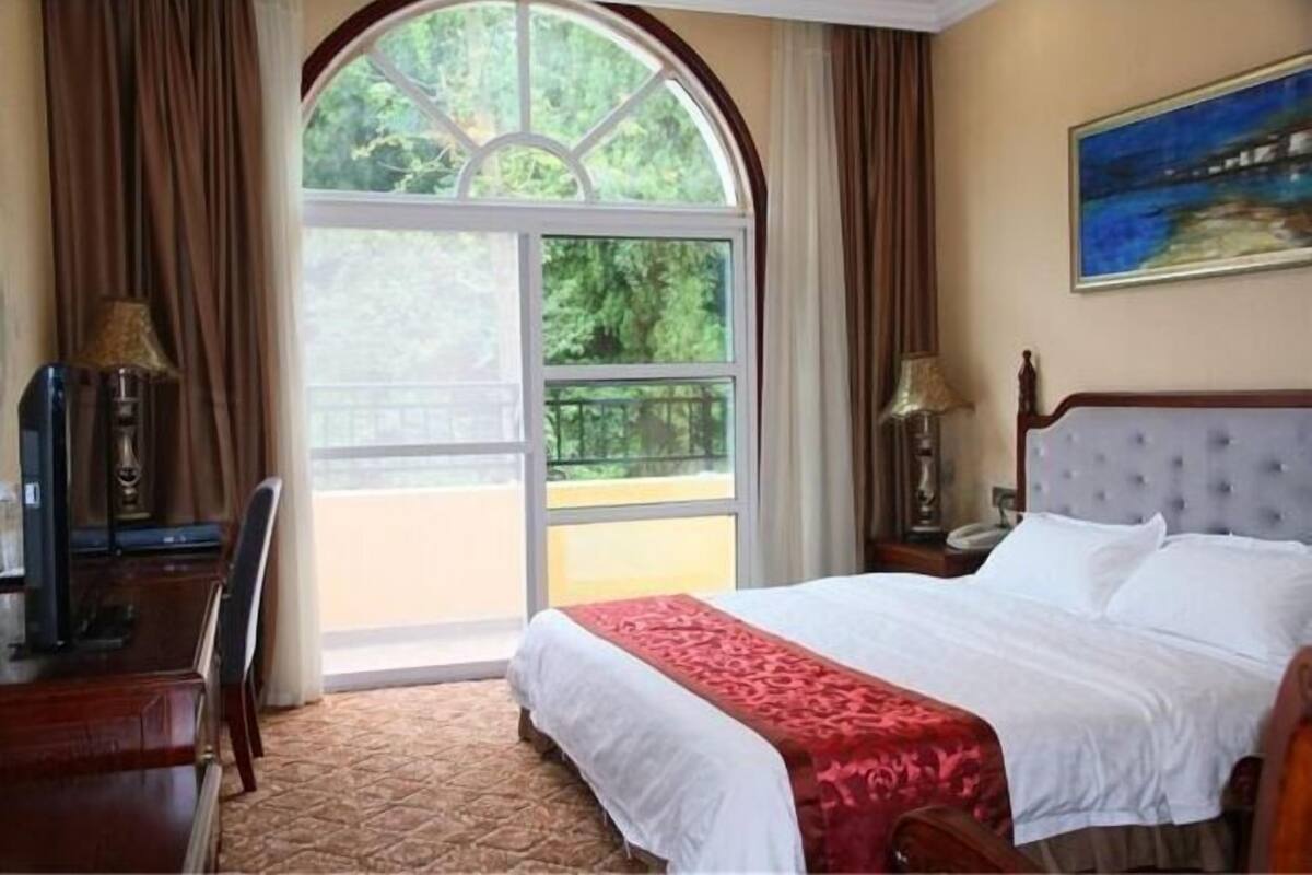 A hotel room with a large window, a bed with white linens, a red throw, a wooden desk, and a painting on the wall.