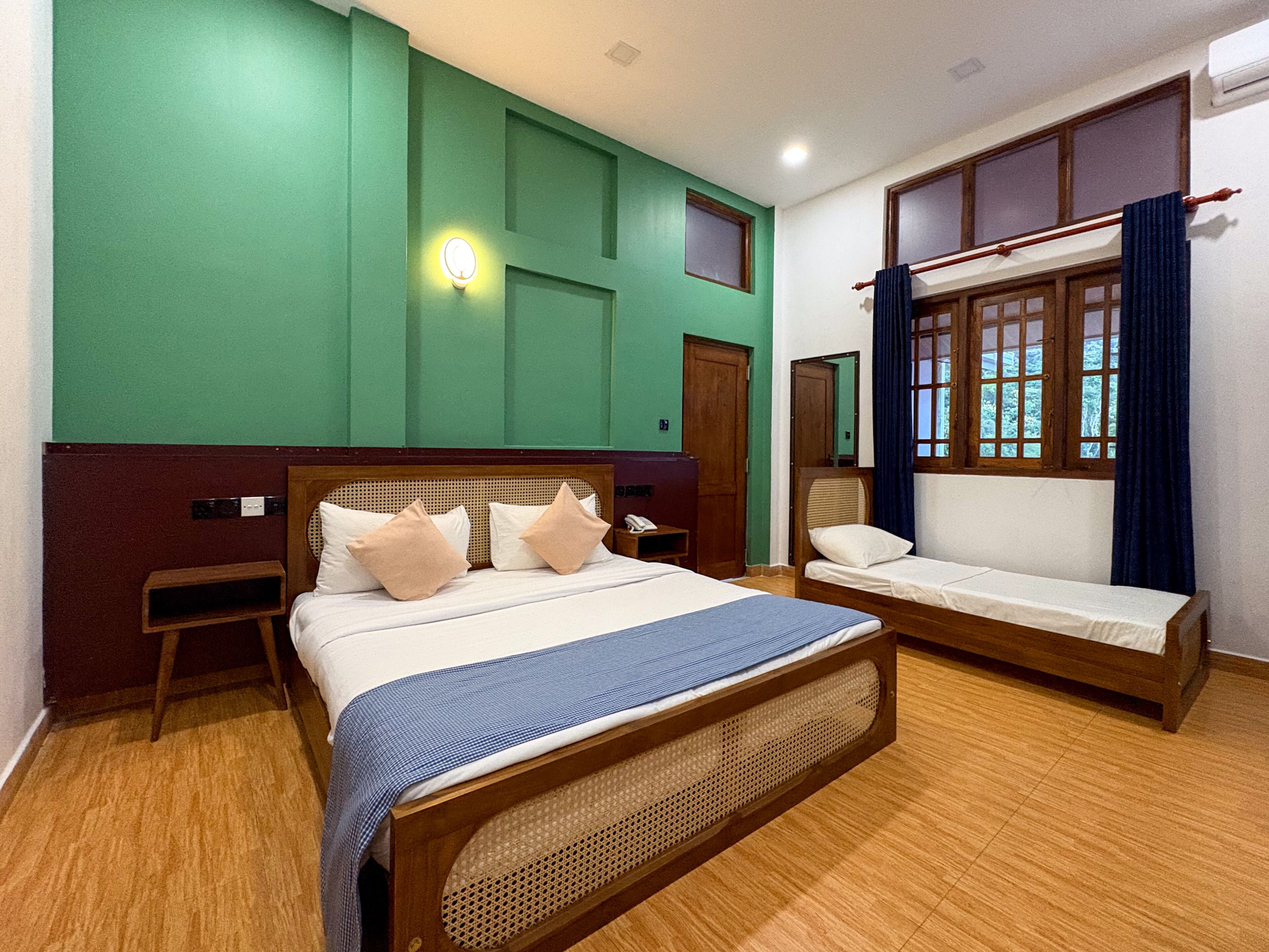 A room with a large bed, a smaller bed, a bedside table, a nightstand, a wall-mounted light, and a window with curtains.