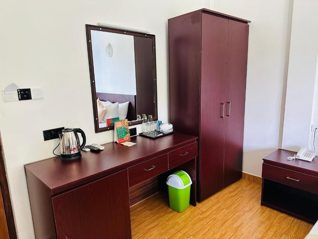Une chambre d’hôtel avec un bureau en bois, un miroir, une armoire, une poubelle et une petite table avec un téléphone.