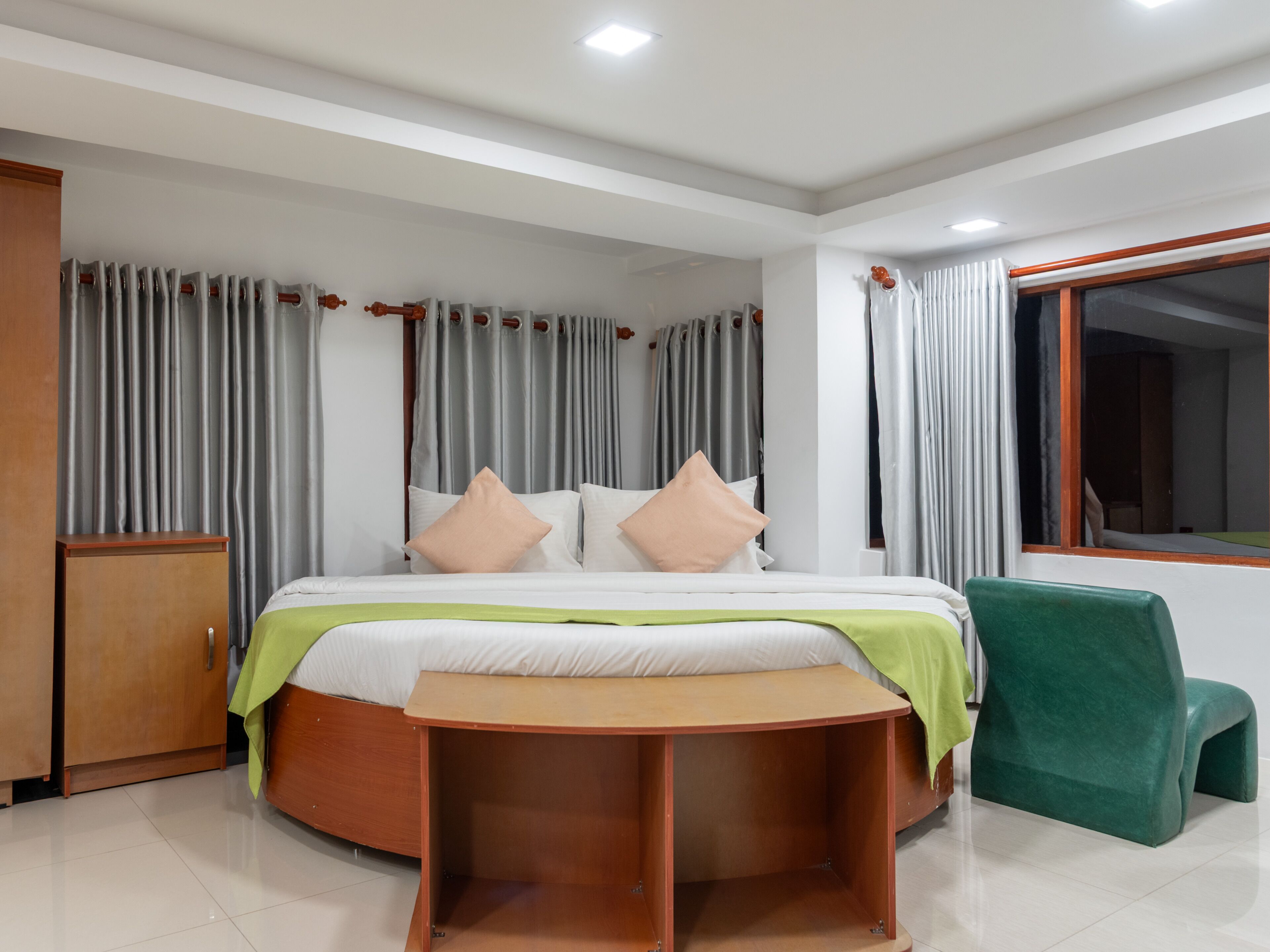 A hotel room with a bed, a wooden nightstand, a green chair, and curtains.